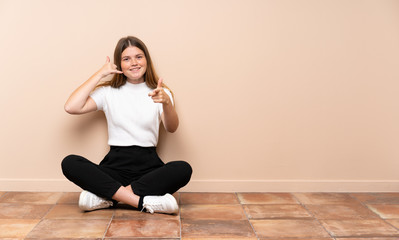 Ukrainian teenager girl sitting on the floor making phone gesture and pointing front