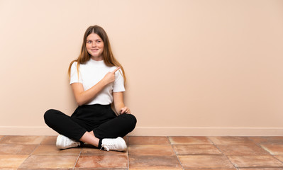 Ukrainian teenager girl sitting on the floor pointing to the side to present a product