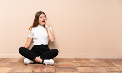 Ukrainian teenager girl sitting on the floor shouting with mouth wide open to the lateral