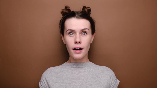 Pretty young woman stares at camera with shocked expression, recieves unexpected news, standing isolated over brown background. Great surprisment or unexpectedness