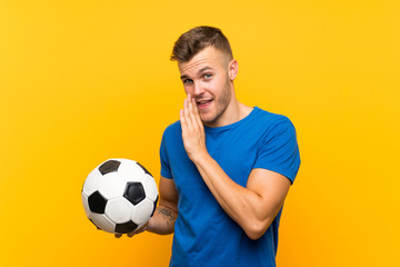 Young handsome blonde man holding a soccer ball over isolated yellow background whispering something