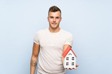 Young handsome blonde man over isolated blue background holding a little house