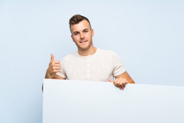 Young handsome blonde man over isolated blue background holding an empty white placard for insert a...