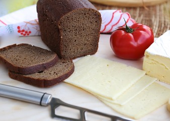Piece of homemade cheese, cut cheese in layers, black bread and knife to cut cheese and greens with tomato on cutting board