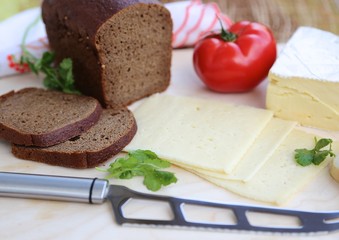 Piece of homemade cheese, cut cheese in layers, black bread and knife to cut cheese and greens with tomato on cutting board