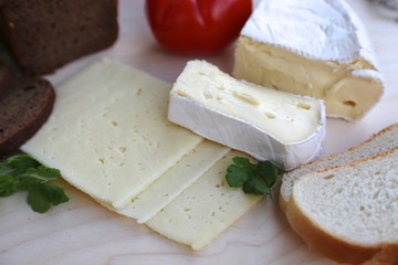 Piece of homemade cheese, cut cheese in layers, black bread and knife to cut cheese and greens with tomato on cutting board