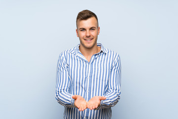 Young handsome blonde man over isolated blue background holding copyspace imaginary on the palm to...
