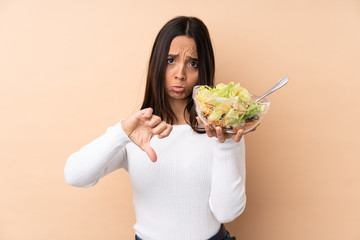 Young brunette girl holding a salad over isolated background showing thumb down with negative expression