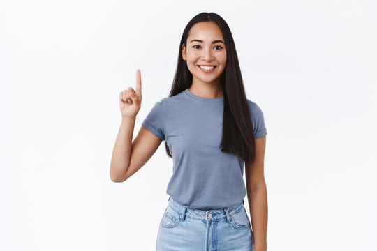 Portrait of cheerful, asian 20s girl in casual t-shirt, pointing fingers up, showing number one, smiling carefree, give advice, promote product, indicating what link click, white background