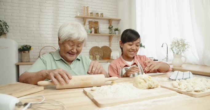 Senior asian woman spending time with her teen granddaughter, cooking together, teaching her how to make dumplings - family ties, generations concept 4k footage