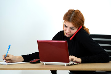 Young office worker woman talking on a cell phone sitting behind working desk with laptop computer and notebook.