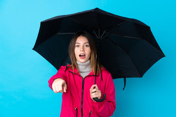 Young woman holding an umbrella over isolated blue wall surprised and pointing front