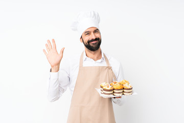 Young man holding muffin cake over isolated white background saluting with hand with happy expression