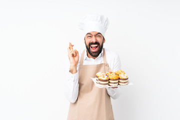 Young man holding muffin cake over isolated white background with fingers crossing