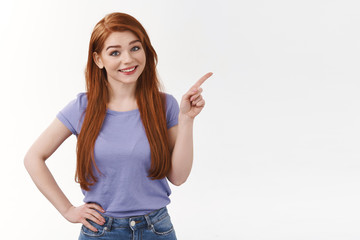 Cute cheerful redhead woman with long ginger hair, pointing upper right corner, tilt head and smiling, invite visit profile, online shop, indicate corporate banner, standing white background