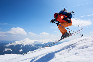 Sportsman skier in skiing equipment jumping in air down steep snowy mountain slope on copy space background of blue sky and highland landscape. Winter risky sports, courage and speed concept.