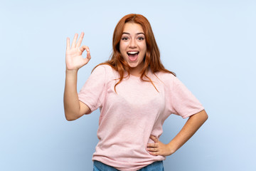 Fototapeta premium Teenager redhead girl over isolated blue background surprised and showing ok sign
