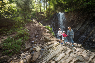 Fototapeta premium Rear view mother and two little daughters are standing on mountain and enjoying bewitching view of waterfall while trekking through mountains on sunny summer day. Concept of tourism with children