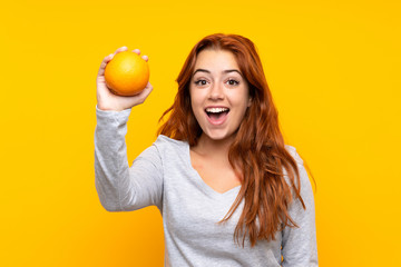 Teenager redhead girl holding an orange over isolated yellow background