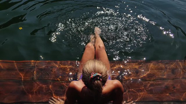 Woman sit on a pier in sunglasses and swimming suit. Girl rest on a flood wood underwater pier. The pavement is covered with water in lake. Lady in the water and splashes feet. View from above.