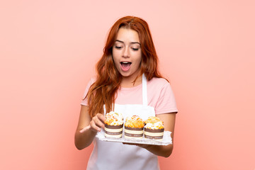 Teenager redhead girl holding lots of different mini cakes over isolated pink background