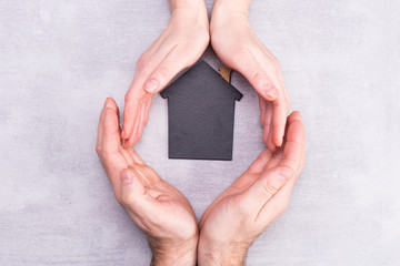 Hands of man and woman surround a model of a dark house on a gray background. Real estate and insurance concept, flat lay, top view