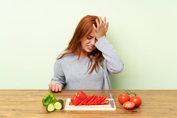 Teenager redhead girl with vegetables in a table having doubts with confuse face expression