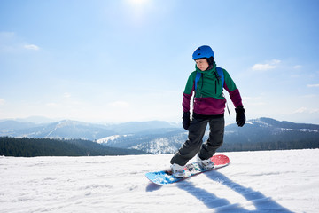 Fototapeta na wymiar Young female snowboarder riding snowboard on copy space background of clear bright blue sky and snowy mountains on sunny winter day. Winter sports and recreation, leisure outdoor activities concept.