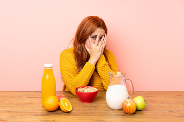 Teenager redhead girl having breakfast in a table covering eyes and looking through fingers