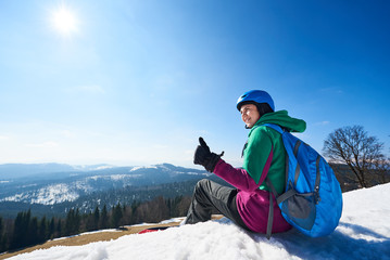 Fototapeta na wymiar Smiling female snowboarder with big backpack resting on snowy summit shows thumb-up gesture on copy space background of blue sky and woody mountains. Extreme winter sports, active lifestyle concept.