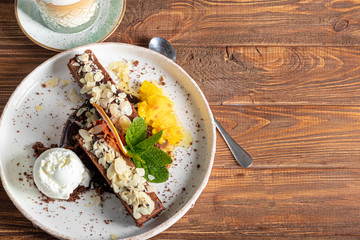 Brownie chocolate dessert with a scoop of white ice cream, slices of mango and a slice of grapefruit. In the background is a cappuccino. On a wooden natural background.