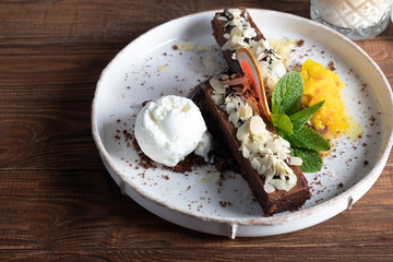 Brownie chocolate dessert with a scoop of white ice cream, slices of mango and a slice of grapefruit. In the background is a cappuccino. On a wooden natural background.