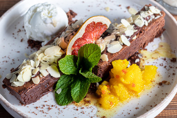 Brownie chocolate dessert with a scoop of white ice cream, slices of mango and a slice of grapefruit. In the background is a cappuccino. On a wooden natural background.