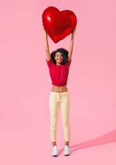 Happy girl holds red heart shape balloon over her head. Photo of smiling young girl in love on pink background. Valentine's Day