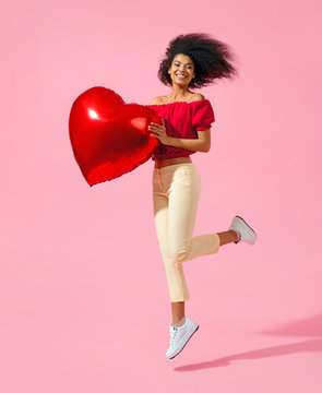Happy girl jumping with red heart shape balloon. Photo of smiling young girl in love on pink background. Valentine's Day