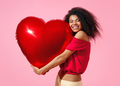 Happy girl holds red heart shape balloon. Photo of smiling young girl in love on pink background. Valentine's Day