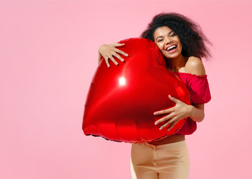 Happy girl holds red heart shape balloon. Photo of smiling young girl in love on pink background. Valentine's Day