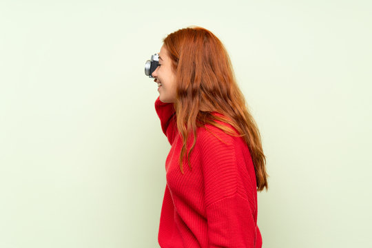 Teenager redhead girl with sweater over isolated green background holding a camera