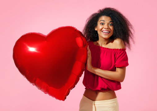 Happy girl holds red heart shape balloon. Photo of smiling young girl in love on pink background. Valentine's Day