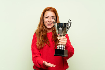 Teenager redhead girl with sweater over isolated green background holding a trophy