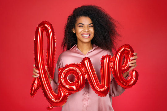 Laughing girl holds letter shaped balloon. Photo of smiling young girl in love on red background. Valentine's Day