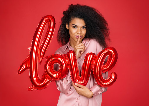 Keep my secret! Beautiful young girl holds forefinger on lips and showing silence gesture. Photo of young girl with letter shaped balloon in love on red background. Valentine's Day