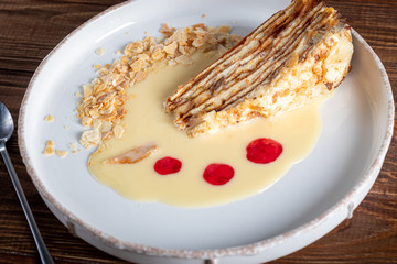 Dessert Napolion with custard, condensed milk sauce, crumbs of dough and raspberry jam. In the background is a cappuccino. On a wooden natural background.