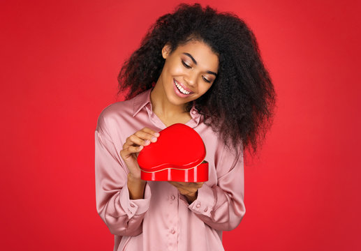 Young excited girl opens gift box. Photo of african american girl in love on red background. Be my Valentine