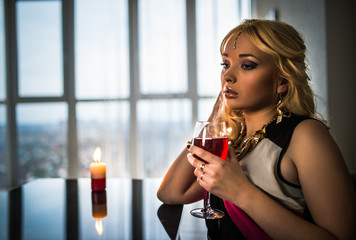 Young pretty blonde woman in jewelry sits at a table with a burning candle and drinks juice or wine on the background of a large window with a city panorama. Concept waiting for a date