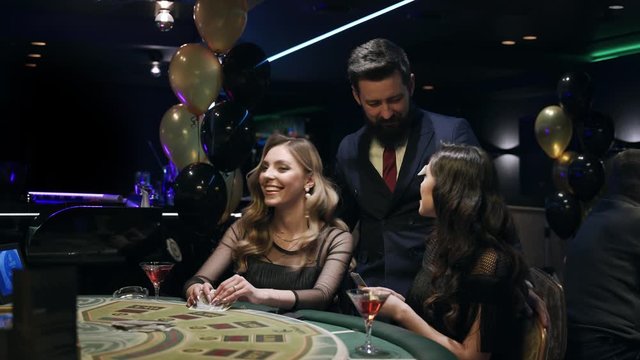 Casino: A bearded man communicates with preatty girls and they smile and laugh. Аlcoholic red champagne in the hands. They are sitting at the pocker table