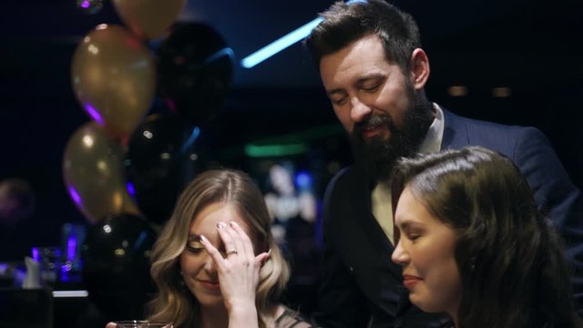 Casino: A bearded man communicates with preatty girls and they smile and laugh. Аlcoholic red champagne in the hands. They are sitting at the pocker table