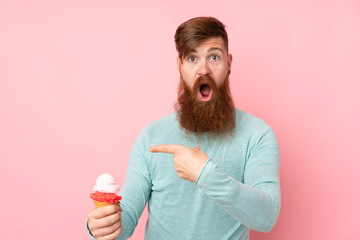 Redhead man with long beard holding a cornet ice cream over isolated pink background surprised and pointing finger to the side