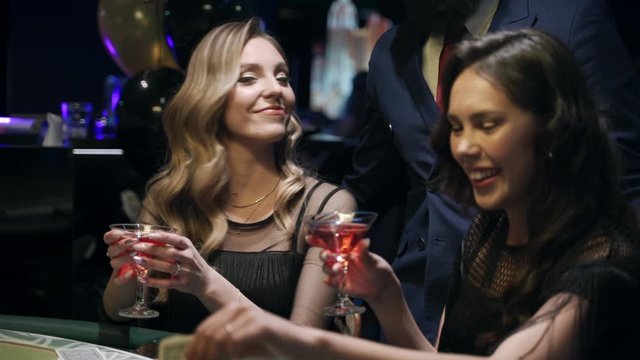 Casino: A bearded man communicates with preatty girls and they smile and laugh. Аlcoholic red champagne in the hands. They are sitting at the pocker table