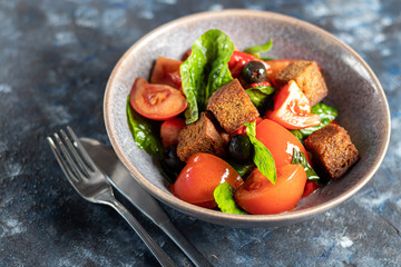 Italian salad of sliced tomatoes, basil leaves, olives, large pieces of croutons. In a deep plate, on a dark background.